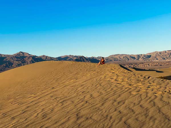 a woman in a red dress sitting at the top of a sand dune looking out at the view at Mesquite San Dunes in Death Valley