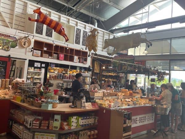 Bustling interior of the Oxbow Market in Napa Valley with a variety of local cheeses on display, customers browsing, and whimsical flying pig sculptures overhead, embodying the region's gourmet food culture.