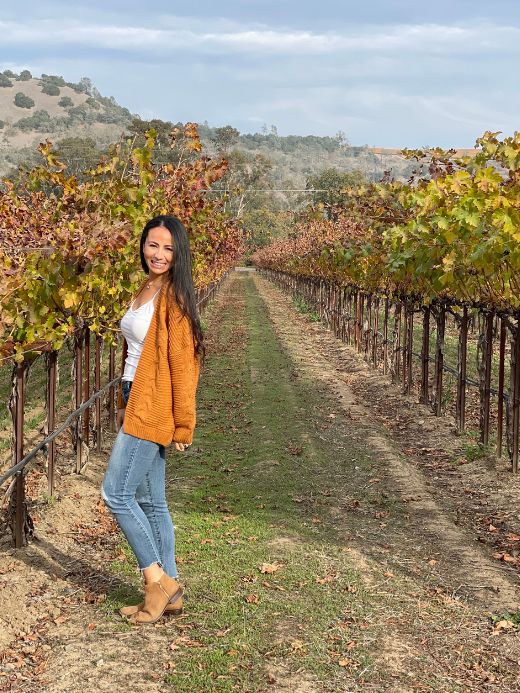 A smiling woman in a cozy mustard cardigan stands among the bare vines of a Napa Valley vineyard in January, capturing the serene beauty of the off-season wine country.