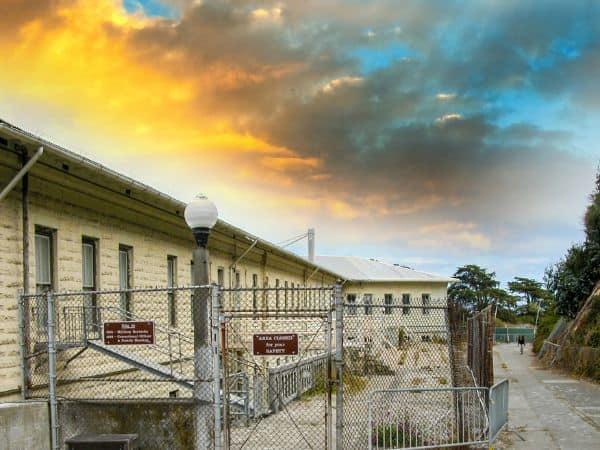 Dramatic sunset skies over Alcatraz Island, with the imposing structure of the prison and warning signs on the fence, evoking the storied past